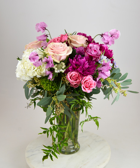 A feminine design in blush pinks and soft whites of roses, peonies, sweet peas, hydrangea, scabiosa with seasonal foliage in a clear tall glass vase.    