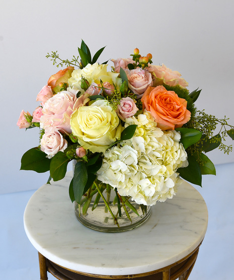 A lush collection of elegant colors of roses with spray roses in hues of peach, pink and white with hydrangea, and seasonal foliage in a glass vase.