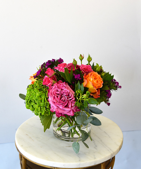 A lush collection of  roses with spray roses in vibrant hues of orange, mauve-pink and pink roses.  Accented with green hydrangea, seasonal blooms and foliage in a glass vase. 