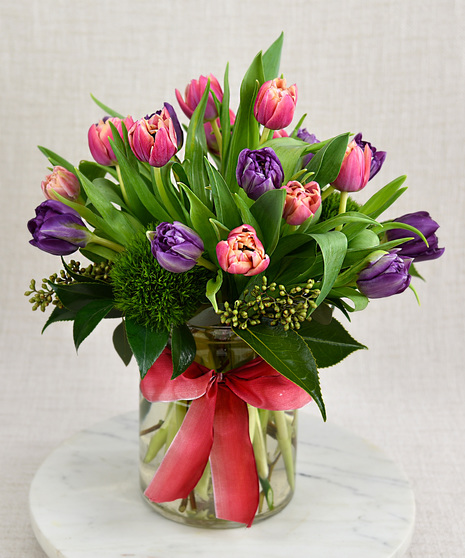 Multi-petal tulips in orange and bright pink or purple. They bloom among seasonal foliage in a glass vase with a spring color ribbon.  