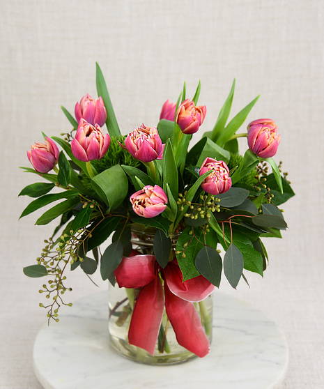 Multi-petal tulips in orange and bright pink or purple. They bloom among seasonal foliage in a glass vase with a spring color ribbon.  